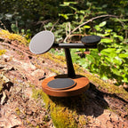 Three-tier black metal and wood wireless charging stand resting on a mossy log outdoors.